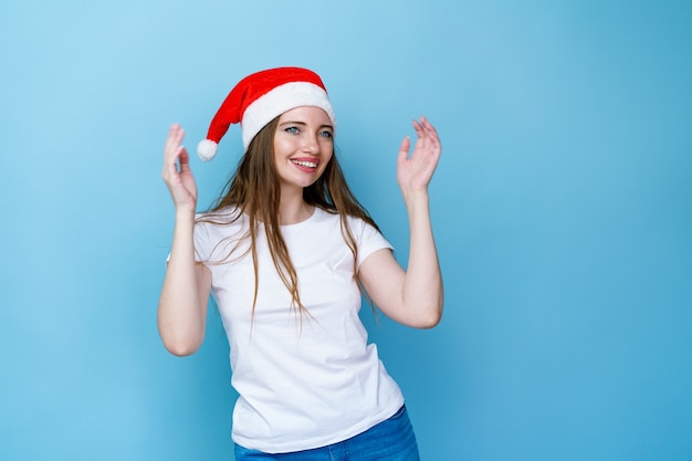 Porträt einer Frau in Weihnachtsmütze und weißem T-Shirt posiert spielerisch auf blauem Hintergrund junger Kaukasier ...