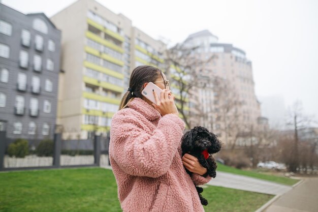 Porträt einer Frau kommuniziert auf dem Smartphone mit einem Welpen in den Händen auf der Straße der Stadt