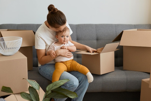 Porträt einer Frau mit Brötchenfrisur, die ein weißes T-Shirt trägt, das auf dem Sofa sitzt und ihr Baby beim Auspacken von Kartons hält, während die junge Familie ihrer Tochter in eine neue Wohnung zieht