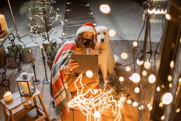 Porträt einer Frau mit ihrem süßen Hund, die Neujahrsfeiertage auf der wunderschön dekorierten Terrasse zu Hause feiert und einen Videoanruf auf einem digitalen Tablet hat