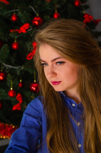 Porträt einer Frau mit leuchtend roten Lippen, blondem langem Haar gegen den Neujahrsbaum. Junge Frau im blauen Herrenhemd. Ferien. Fröhliche Weihnachten. lustiges Gesicht