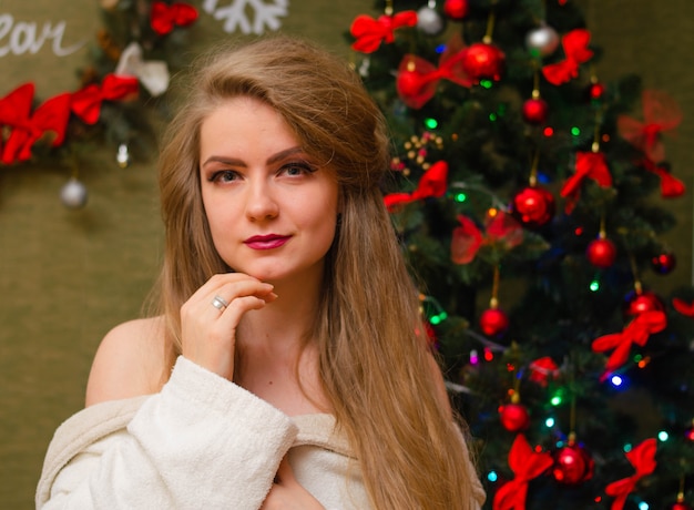 Porträt einer Frau mit leuchtend roten Lippen, blondem langem Haar gegen den Neujahrsbaum. Junge Frau in einem weißen warmen Mantel, Schultern sind sichtbar. Ferien. Fröhliche Weihnachten
