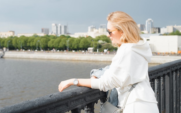Porträt einer Frau mit Sonnenbrille, die auf der Brücke steht und den Damm und den Fluss betrachtet. Junge Blondine in lässiger, leichter Kleidung bei einem Spaziergang in der Stadt. Reisetourismus an einem sonnigen Sommertag.