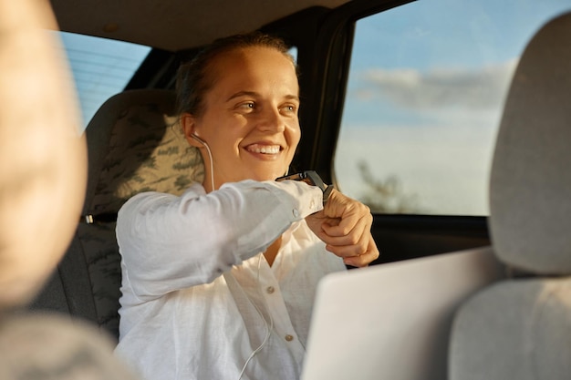 Porträt einer Frau mit weißem Hemd, die auf dem Rücksitz des Autos sitzt und mit einem Laptop unter Verwendung einer intelligenten Uhr arbeitet, um eine Sprachnachricht aufzuzeichnen, die glücklich lächelt