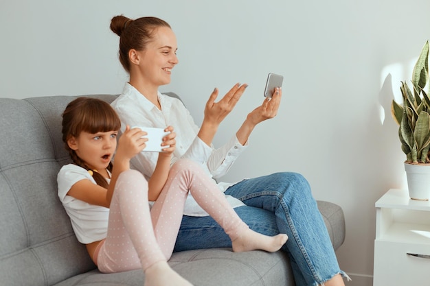 Porträt einer Frau mit weißem Hemd und Jeans, die mit ihrer Tochter auf dem Sofa sitzt und ein Smartphone hält und einen Videoanruf hat, Kind, das Spiele über Mobiltelefon mit schockiertem Ausdruck spielt.