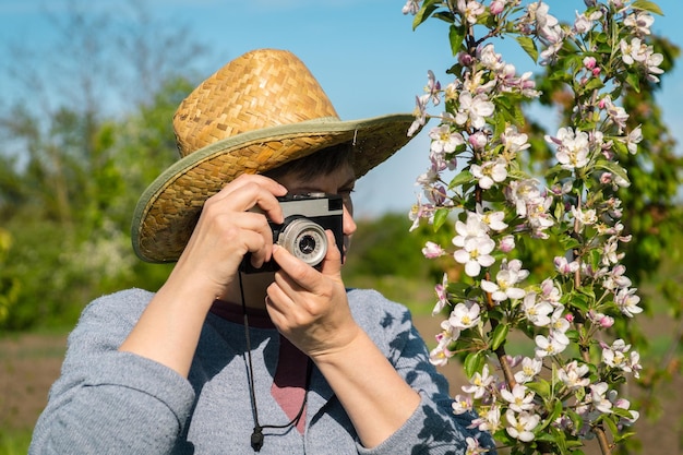 Porträt einer Frau mittleren Alters in einem Strohhut mit einer alten Kamera, die einen blühenden Zweig fotografiert