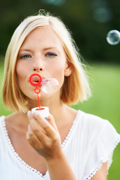 Porträt einer Frau und Blasen in der Natur für einen lustigen Tag oder beim Spielen im Outdoor-Park Gesicht einer jungen glücklichen Frau oder Blondine mit einem Stock oder einem Zauberstab, um Blasen in einem spielerischen Urlaub oder in einer Sommerpause zu blasen