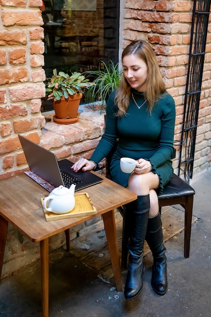 Porträt einer Freiberuflerin der jungen Frau, die Laptop-Computer für Fernarbeit beim Sitzen im modernen Kaffee mit Tasse Tee verwendet