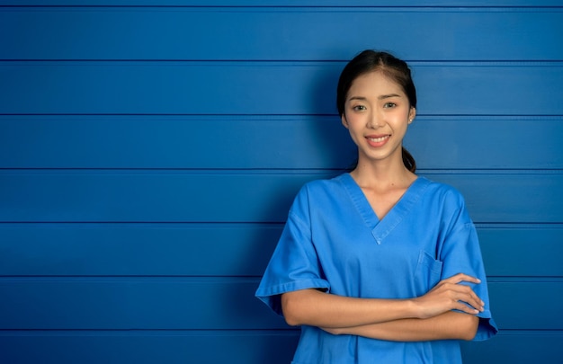 Porträt einer freundlichen, fröhlich lächelnden, selbstbewussten asiatischen Ärztin oder Krankenschwester im blauen Anzug stehen mit verschränkten Armen auf Holzwand Hintergrund medizinisches Fachpersonal in blauen Scrubs mit Kopierraum