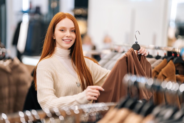 Porträt einer freundlichen, hübschen jungen Kundin, die Kleidung aus dem Regal im Bekleidungsgeschäft auswählt