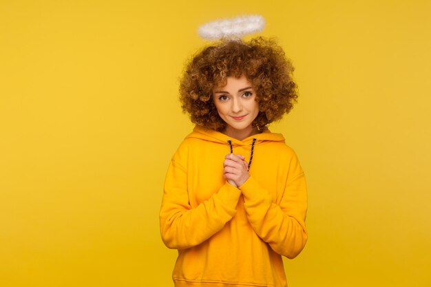 Porträt einer freundlichen, lockigen Hipster-Frau mit Heiligem Nimbus, die Hände im Gebet hält und mit gehorsamem, bescheidenem Ausdruck in die Kamera schaut. Studioaufnahme im Innenbereich isoliert auf gelbem Hintergrund