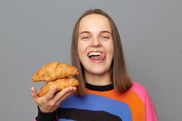 Porträt einer fröhlich lächelnden kaukasischen Frau mit Pullover, die zwei Croissants in den Händen hält und in die Kamera schaut, die die Zunge herausstreckt und isoliert vor grauem Hintergrund posiert