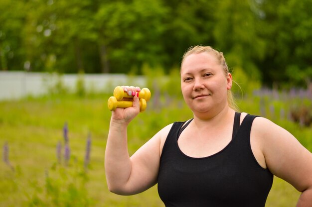 Porträt einer fröhlichen alten Frau in Fitnesskleidung, die mit gelben Hanteln im Park trainiert