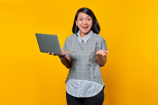 Porträt einer fröhlichen asiatischen Frau mit Brille, die Laptop hält und die Hand auf gelbem Hintergrund hebt