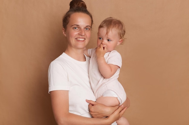 Porträt einer fröhlichen Frau mit Brötchenfrisur, die ein weißes T-Shirt trägt und mit ihrer kleinen Tochter in den Händen steht, isoliert vor braunem Hintergrund, die mit einem zahnigen Lächeln in die Kamera blickt