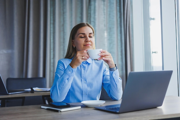 Porträt einer fröhlichen Frau mit klassischer Brille, die in der Freizeit im Café mit Kaffee lächelt positive europäische Frau im blauen Hemdschreibtisch mit Laptop-Fernarbeit