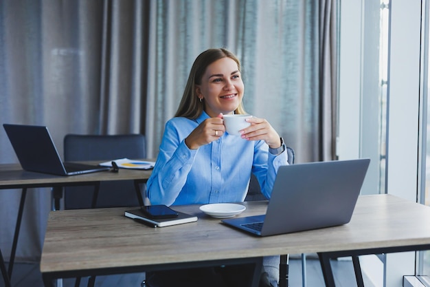 Porträt einer fröhlichen Frau mit klassischer Brille, die in der Freizeit im Café mit Kaffee lächelt positive europäische Frau im blauen Hemdschreibtisch mit Laptop-Fernarbeit