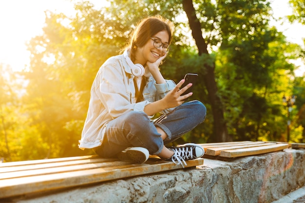 Porträt einer fröhlichen, fröhlichen, süßen jungen Studentin mit Brille, die draußen im Naturpark mit Handy sitzt.