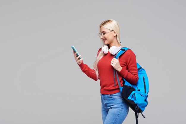 Porträt einer fröhlichen glücklichen blonden Studentin mit blauem Rucksack, die Online-Vortrag am Telefon mit Kopfhörern hört, die über grauem Raum isoliert werden. Kopieren Sie den Platz für Text auf der linken Seite