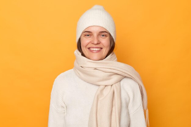 Porträt einer fröhlichen, glücklichen jungen erwachsenen frau mit weißer pullovermütze und schal, die vor der gelben wand posiert und in guter laune lächelt und glücklich positive emotionen ausdrückt