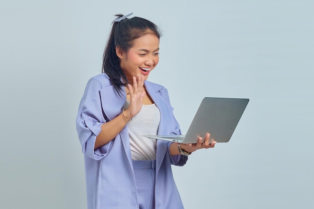 Porträt einer fröhlichen jungen asiatischen Frau, die dem Laptop isoliert auf weißem Hintergrund eine Hand winkt