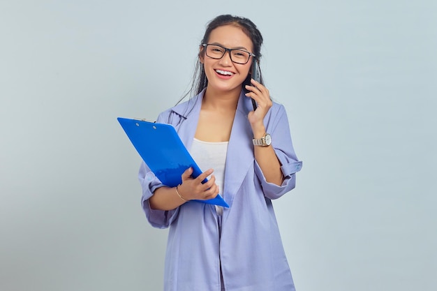 Porträt einer fröhlichen jungen asiatischen Frau, die lacht, während sie am Handy mit Freunden über etwas Lustiges spricht, während sie einen Dokumentenordner isoliert auf violettem Hintergrund hält