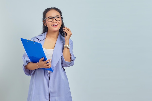 Porträt einer fröhlichen jungen asiatischen Frau, die lacht, während sie am Handy mit Freunden über etwas Lustiges spricht, während sie einen Dokumentenordner isoliert auf violettem Hintergrund hält