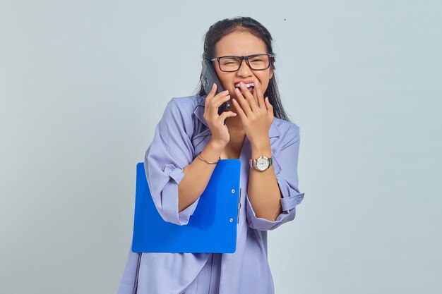 Porträt einer fröhlichen jungen asiatischen Frau, die lacht, während sie am Handy mit Freunden über etwas Lustiges spricht, während sie einen Dokumentenordner isoliert auf violettem Hintergrund hält