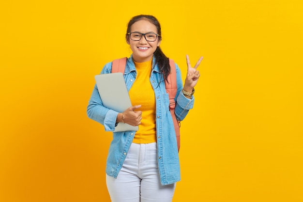 Porträt einer fröhlichen jungen asiatischen Studentin in Freizeitkleidung mit Rucksack, der Laptop hält und Friedenszeichen mit dem Finger zeigt, isoliert auf gelbem Hintergrund Bildung im Universitätskolleg-Konzept