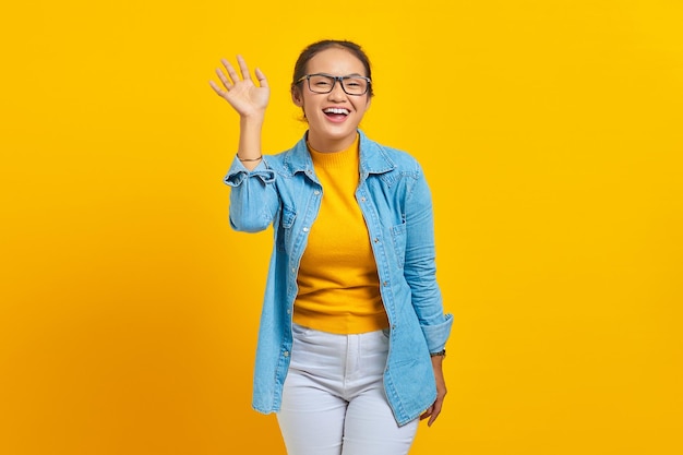 Porträt einer fröhlichen jungen asiatischen Studentin in Jeanskleidung, die mit der Hand in die Kamera winkt, isoliert auf gelbem Hintergrund. Menschen aufrichtige Emotionen Lifestyle-Konzept