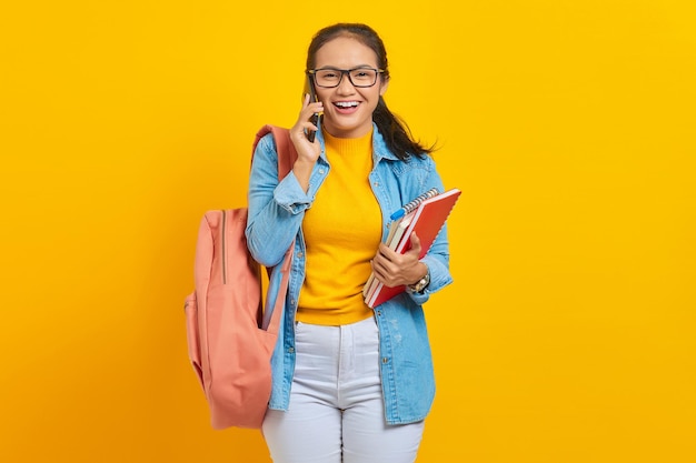 Porträt einer fröhlichen jungen asiatischen Studentin in Jeanskleidung mit Rucksack, die am Handy spricht und Buch isoliert auf gelbem Hintergrund hält