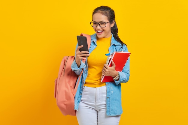 Porträt einer fröhlichen jungen asiatischen Studentin in Jeanskleidung mit Rucksack mit Handy, die SMS-Nachricht tippt und Bücher isoliert auf gelbem Hintergrund hält