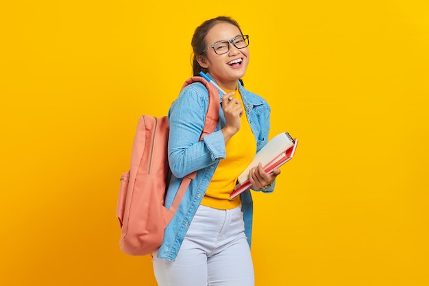 Porträt einer fröhlichen jungen asiatischen Studentin in lässiger Kleidung mit Rucksack, der Buch und Stift hält und auf die Kamera blickt, die auf gelbem Hintergrund isoliert ist Bildung im College-Universitätskonzept