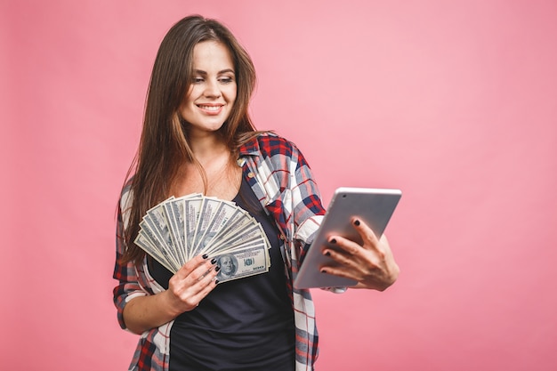Porträt einer fröhlichen jungen Frau, die Geldbanknoten hält und lokalisiert über rosa Hintergrund feiert. Tablet verwenden.
