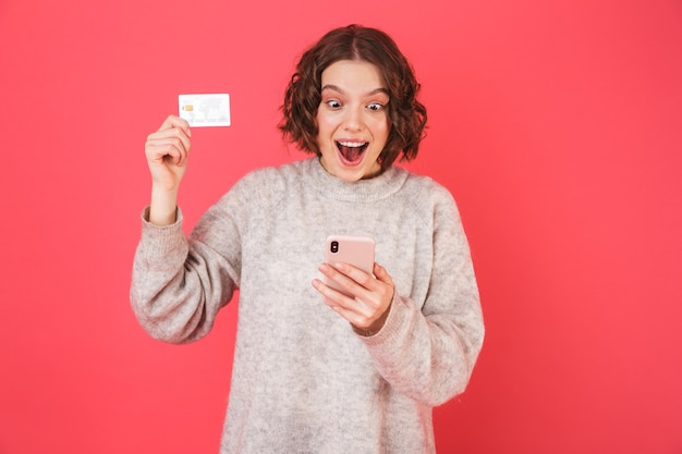 Porträt einer fröhlichen jungen Frau, die isoliert über rosa steht, unter Verwendung des Mobiltelefons, das Plastikkreditkarte zeigt