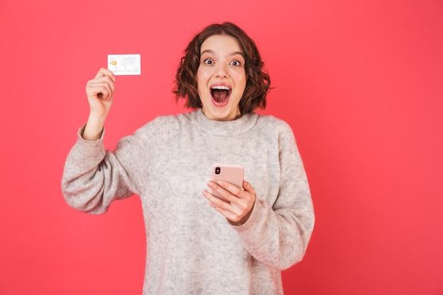 Porträt einer fröhlichen jungen Frau, die isoliert über rosa steht, unter Verwendung des Mobiltelefons, das Plastikkreditkarte zeigt