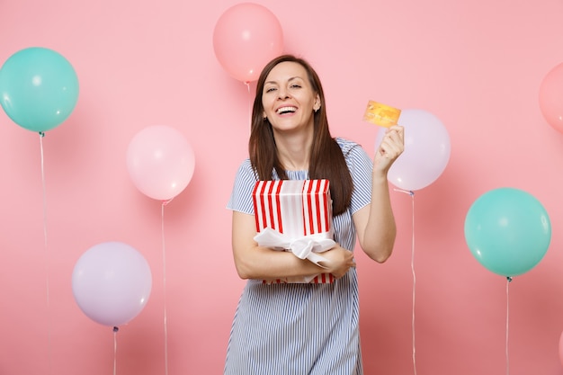 Porträt einer fröhlichen jungen Frau im blauen Kleid mit Kreditkarte und roter Schachtel mit Geschenk auf pastellrosa Hintergrund mit bunten Luftballons. Geburtstagsfeier, Menschen aufrichtige Emotionen.