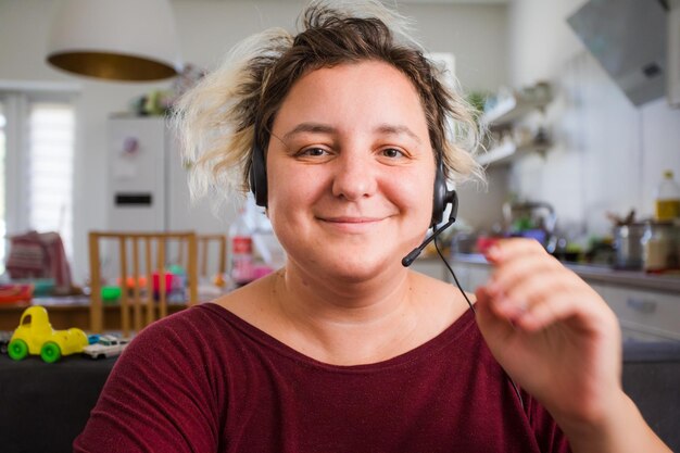 Porträt einer fröhlichen jungen Frau im Headset während einer Online-Konferenz Junge Mutter, die von zu Hause aus arbeitet und Kinder großzieht Leichte Küche und Sofa mit kleinen Spielsachen im Hintergrund