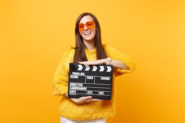 Porträt einer fröhlichen jungen Frau in Pelzpullover mit orangefarbener Herzbrille, die klassische schwarze Filmklappe hält, die auf gelbem Hintergrund isoliert ist. Menschen aufrichtige Emotionen Lebensstil. Werbefläche.