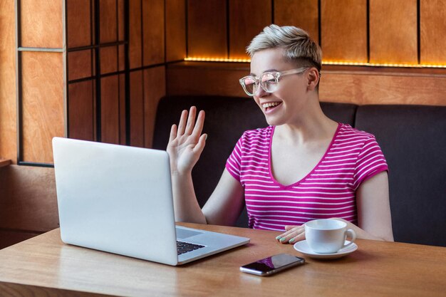 Porträt einer fröhlichen jungen frau mit kurzen haaren in rosa t-shirt und brille sitzt im café und spricht mit ihrer freundin auf dem laptop und zeigt der webcamera ein hallo-zeichen mit einem zahnigen lächeln, drinnen