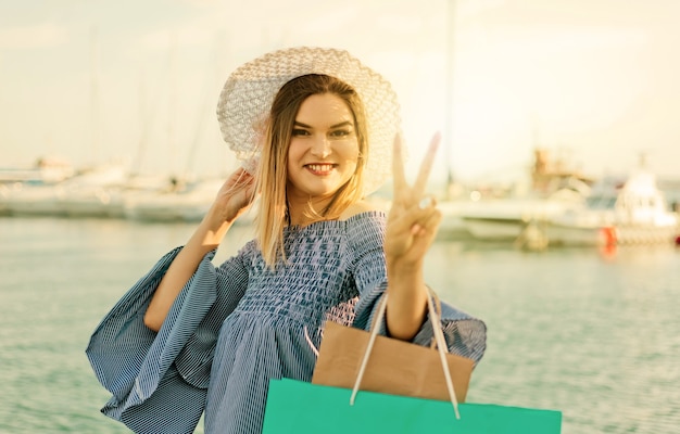 Porträt einer fröhlichen jungen Shopaholic-Frau in Hut und Kleid auf dem Hintergrund des Meeres. Glückliches Einkaufskonzept.