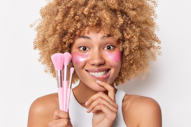 Porträt einer fröhlichen, neugierigen Frau, die Lippen beißt, sieht glücklich aus, trägt rosa Hydrogelpflaster unter den Augen auf, um Falten zu reduzieren, hält Kosmetikpinsel, steht mit nackten Schultern vor weißem Hintergrund