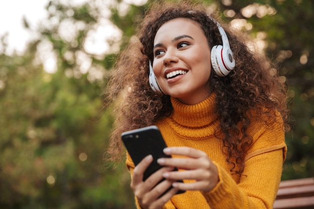 Porträt einer fröhlichen optimistischen schönen jungen lockigen Frau sitzen auf der Bank im Park im Freien und hören Musik mit Kopfhörern unter Verwendung des Mobiltelefons.