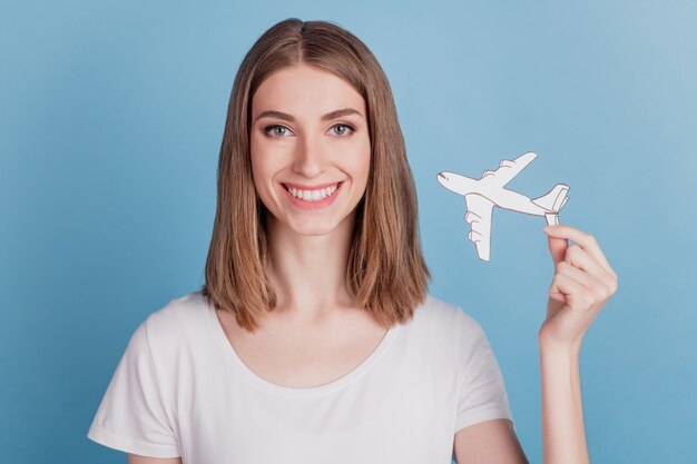 Porträt einer fröhlichen positiven Dame, die weißes Papierflugzeug mit einem strahlenden Lächeln auf blauem Hintergrund hält