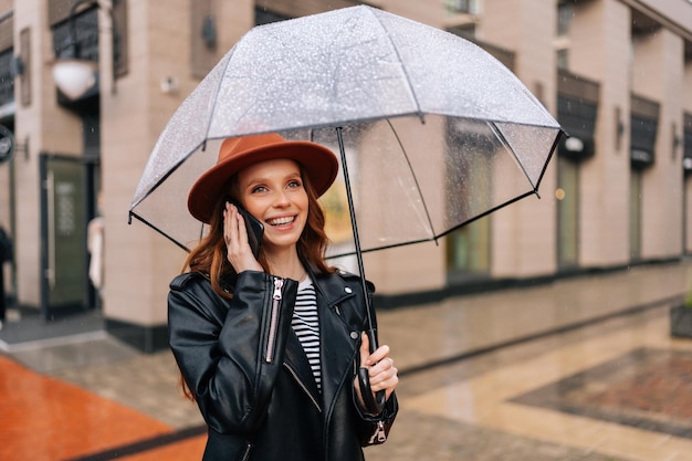 Porträt einer fröhlichen rothaarigen jungen Frau mit modischem Hut, die mit Regenschirm steht und im Regen auf der Straße der europäischen Stadt mit dem Handy spricht