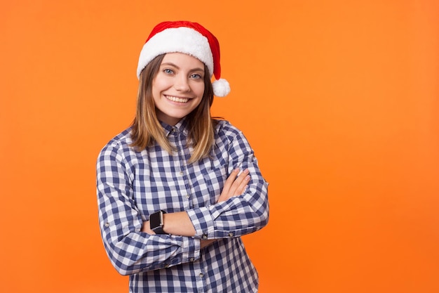 Porträt einer fröhlichen, schönen, brünetten Frau mit Weihnachtsmütze und kariertem Hemd, die mit verschränkten Armen steht und fröhlich in die Kamera blickt, festliche Stimmung, Studioaufnahme isoliert auf orangefarbenem Hintergrund