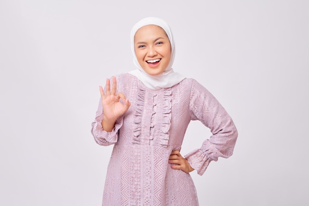 Porträt einer fröhlichen, schönen jungen asiatischen muslimischen Frau mit Hijab und lila Kleid, die ein Okay-Zeichen zeigt, isoliert auf weißem Studiohintergrund