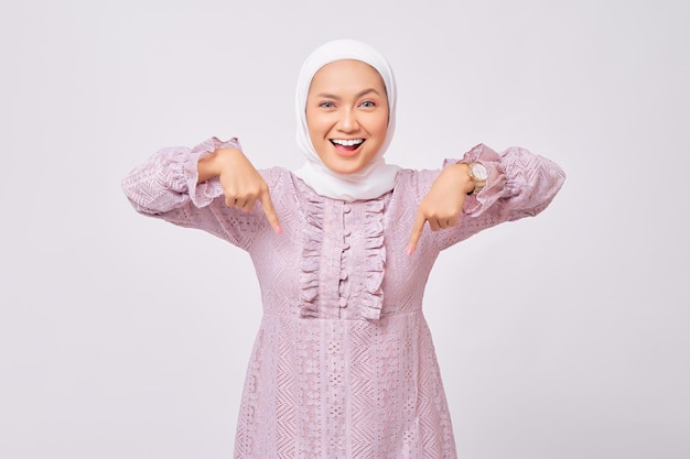 Porträt einer fröhlichen, schönen jungen asiatischen muslimischen Frau mit Hijab und lila Kleid, die mit dem Finger nach unten zeigt und in die Kamera schaut, isoliert auf weißem Studiohintergrund