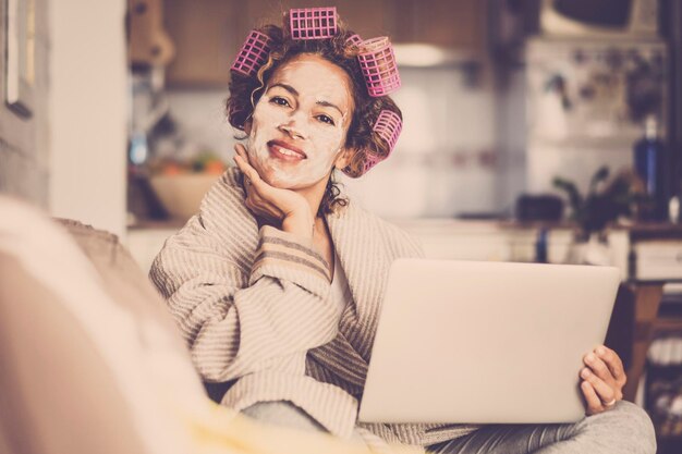 Porträt einer fröhlichen und glücklichen jungen Frau, die mit Schönheitsbehandlungs-Gesichtscreme und rosa Lockenwicklern auf dem Haar lächelt und in die Kamera schaut Konzept der Pflege selbst weiblicher Menschen zu Hause auf dem Sofa Laptop