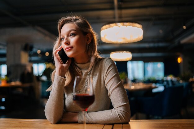 Porträt einer gelangweilten hübschen jungen Frau, die auf dem Smartphone mit einem Freund spricht, der spät mit einem Glas Rotwein im Restaurant sitzt